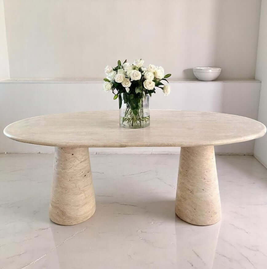 Stylish oval travertine dining table with a vase of white roses, showcasing natural elegance for home decor.